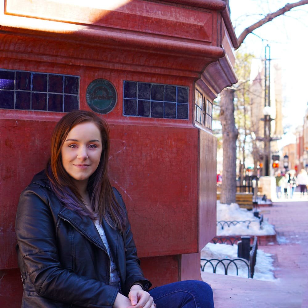 A woman with long brown hair in a black jacket sits by a red tiled structure on a sunny city sidewalk with snow in the background.